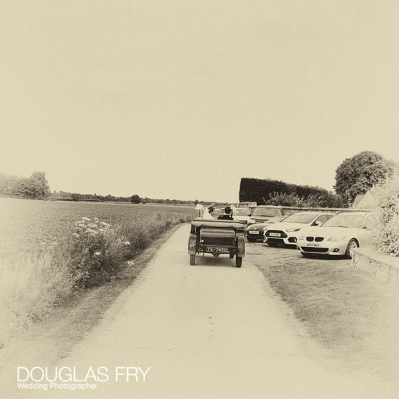 Couple driving to reception across field in vintage car